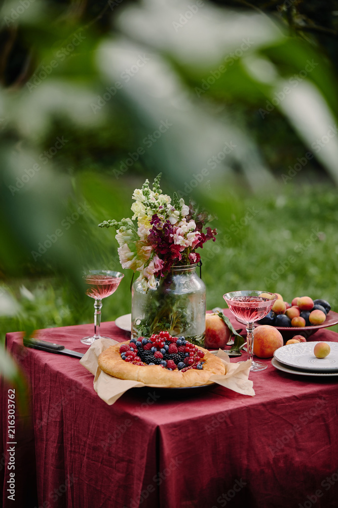 bouquet of flowers in glass jar, berries pie and wineglasses on table in garden