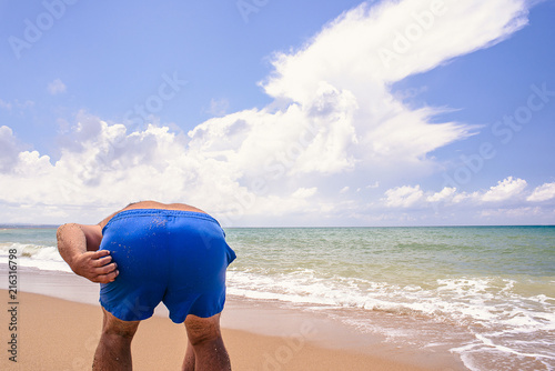 Fotografie fat man stood in front of the camera back and bent down, spoiling the seascape with his ass
