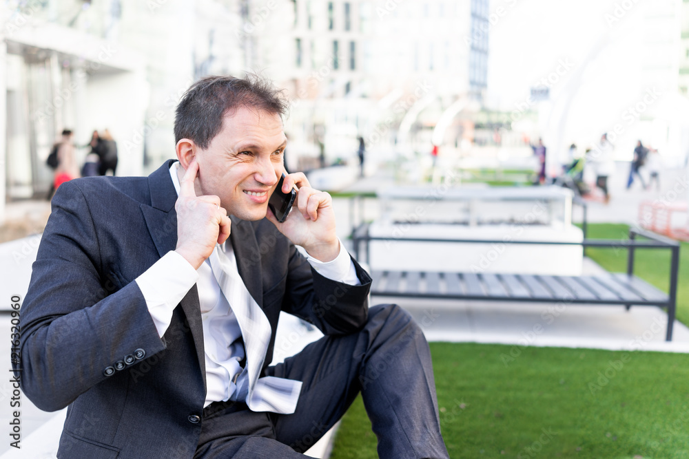 Funny businessman sitting on bench making face hearing impaired talking holding smartphone phone mobile cellphone in suit and tie on break in park