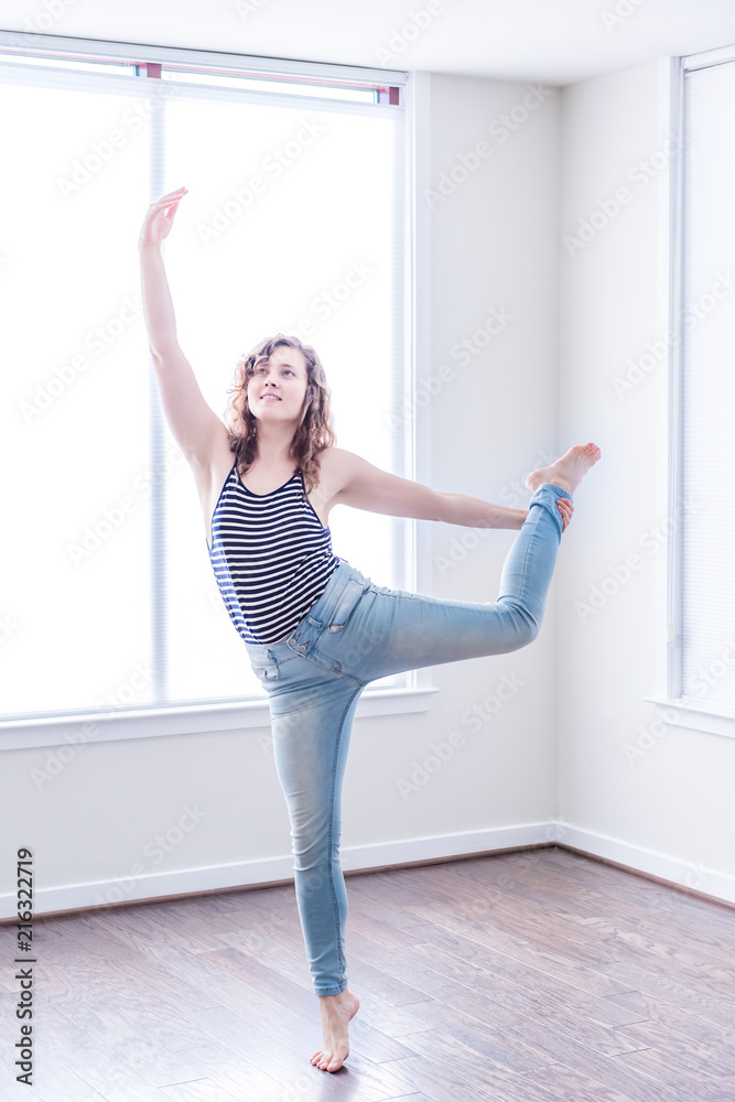 Young happy woman stretching dancing in empty modern new room with hardwood floors and large sunny windows in apartment