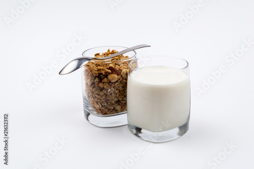 A glass full of milk and a glass full of muesli with a stainless steel spoon on a white background