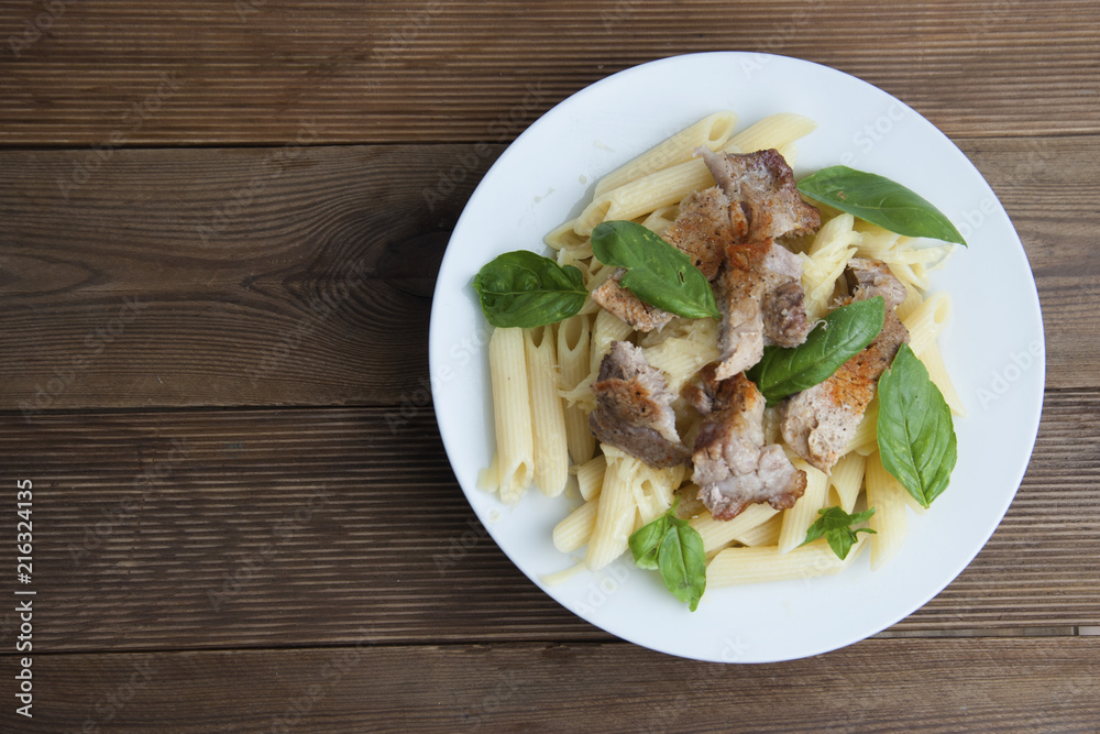 Cooked penne pasta isolated in white round plate, with basil leaves and pork meat, over wooden table, decorated with basil leaves. Top view. Cooked penne pasta isolated in white round plate, with basil leaves and pork meat, over wooden table, decorated with basil leaves. Top view.