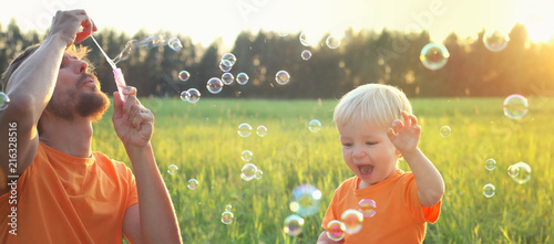 Cute toddler blond boy and his father playing with soap bubbles on summer field. Beautiful sunset light. Happy childhood concept. Authentic lifestyle image