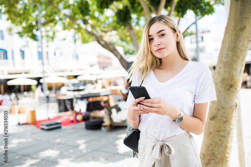 Young blonde woman walking and typing on phone in the street in a sunny summer day