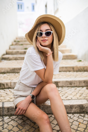 Happy young blonde woman in hat and sunglasses sitting on stairs