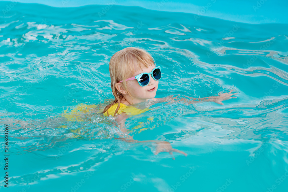Little girl in a yellow swimsuit and sunglasses is swimming and jumping to swimming pool at resort. Summer fun in the pool. 