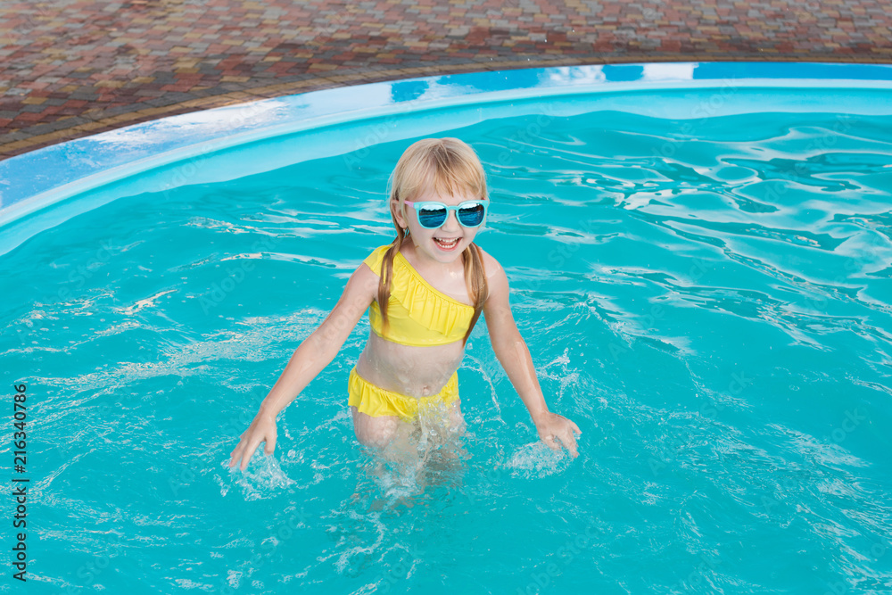 Little girl in a yellow swimsuit and sunglasses is swimming and jumping to swimming pool at resort. Summer fun in the pool. 