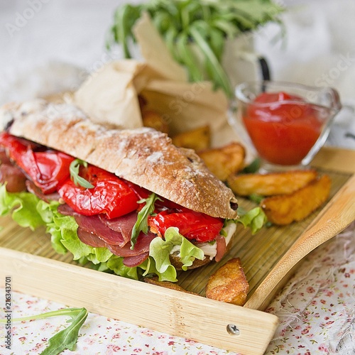 sandwich with ham, cream cheese, lettuce leaves, arugula, red pepper, baked potatoes, ketchup on a white wooden background with a floral fabric