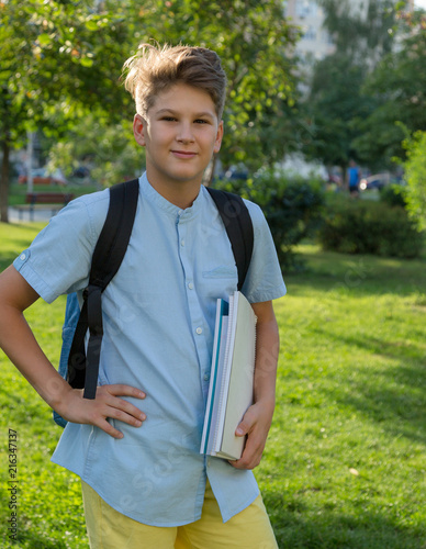 cute, young schoolboy in blue shirt with backpack and workbooks stands in the park. Back to school, education concept.  © Natali