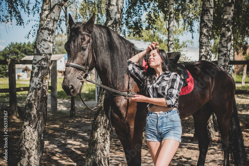 Woman enjoying horse company. Young Beautiful Woman dressed plaid shirt With black Horse Outdoors, stylish girl at american country style 