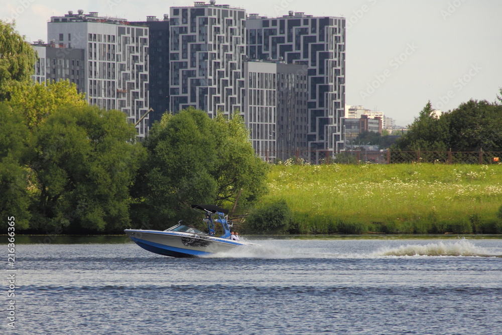 Obraz premium Silver-blue towing boat fast floating on the river in summer on the background of water, shore with green trees and modern city building in Moscow, Strogino