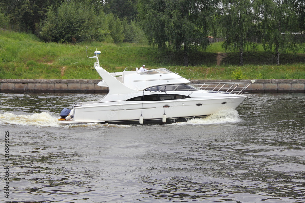 Travel, luxury water recreation on boat - a white motor yacht with radar on the targa slowly sails along the concrete shores of the navigable canal in summer amid green trees on the shore, side view