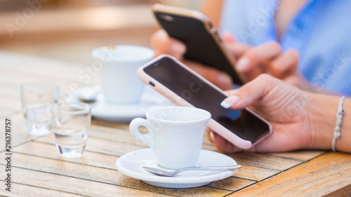 Close-up of female hands holding smartphones chatting on social network or working in cafe with two cups of italian espresso coffe end two little glasses of water, focus on the first cup of coffee.