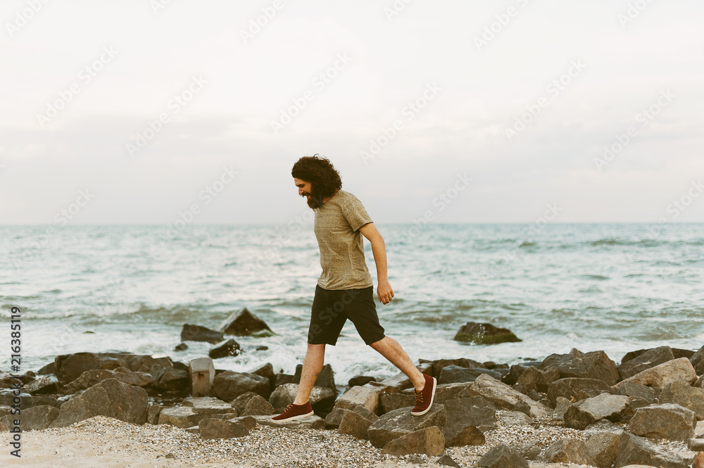 Young bearded hipster man walking on the beach stones looking down.