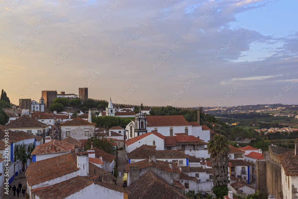 View of narrow streets and castle of Obidos from wall of fortress. Scenic old town with medieval architecture in the sunset. White houses red tiled roofs.