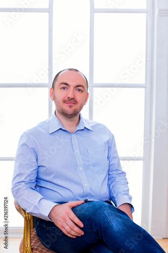 Portrait of stylish man in blue shirt, sits in an armchair on background window 
