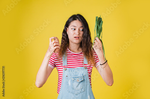 young girl in denim overalls and striped t-shirt holding potatoes and onions on a yellow isolated background