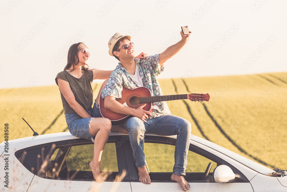 Beautiful young couple taking self portrait from car roof with smart phone against yellow field background.