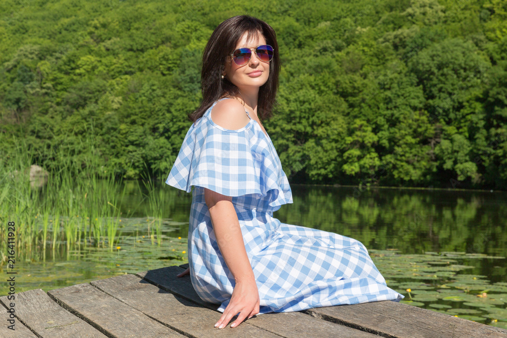 Naklejka premium Young woman in sunglasses sits on the bridge near the river on a sunny day.