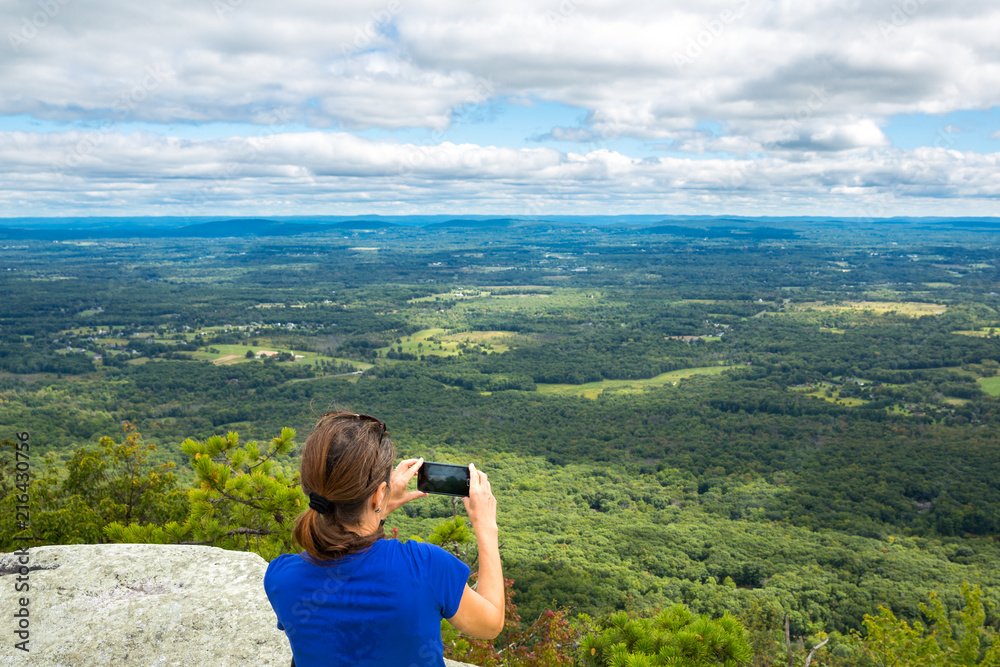 Woman takes a snpashot of the Hudson Valley farm land from Gertrude's Nose hiking trail, in Upstate New York Woman takes a snpashot of the Hudson Valley farm land from Gertrude's Nose hiking trail, in Upstate New York