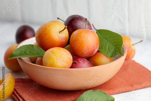 Fototapeta Naklejka Na Ścianę i Meble -  Fresh colorful plums with leaves in fruit bowl on white stone background, close up view