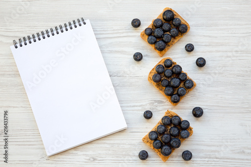 Vegan toasts with peanut butter, blueberries and chia seeds on a white wooden background, top view. Healthy eating. Flat lay, from above. Blank notebook.