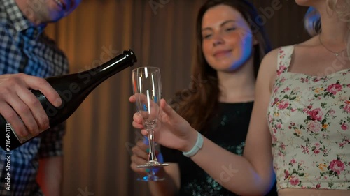Handsome man pouring champagne white sparkled wine in glasses of two attractive sexy girls in evening dresses. Close-up. Comfortable party at apartment. 4k UltraHD. photo