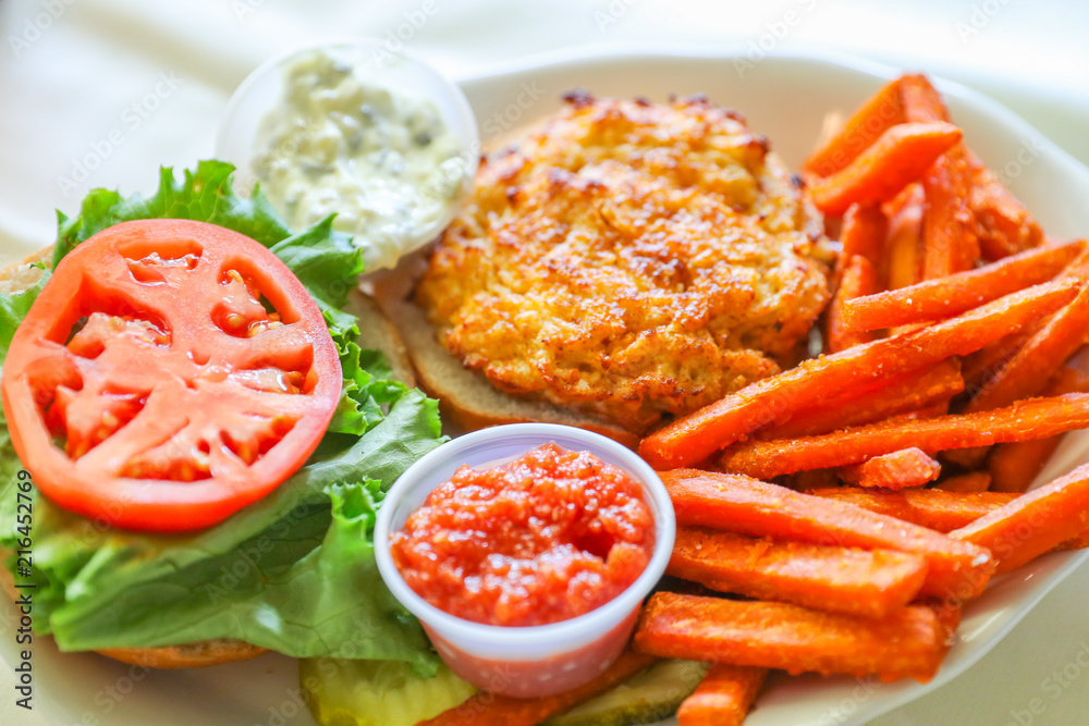 A hot, fresh, crab cake sandwich with tomato slices and sweet potato fries