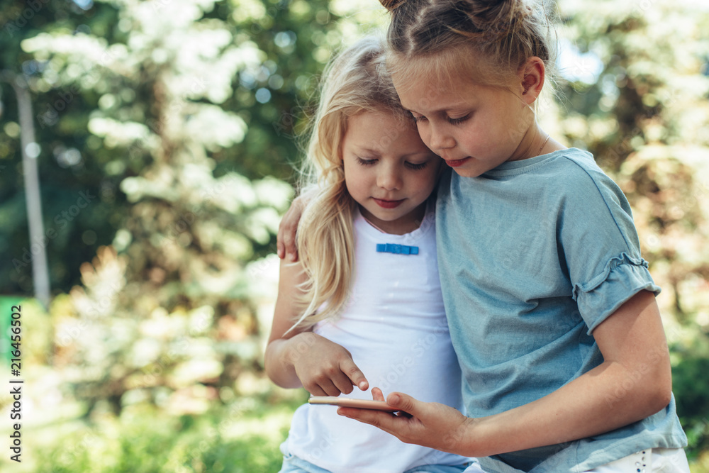 Waist up portrait of small girls standing in cuddles with phone in hands. They are playing games on smartphone peacefully. Copy space in left side Waist up portrait of small girls standing in cuddles with phone in hands. They are playing games on smartphone peacefully. Copy space in left side
