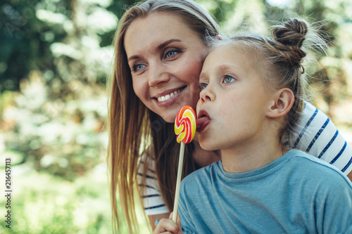 Waist up portrait of smiling woman bonding to kid in park. Small child is licking lollypop with pleasure. They are admiring leisure together