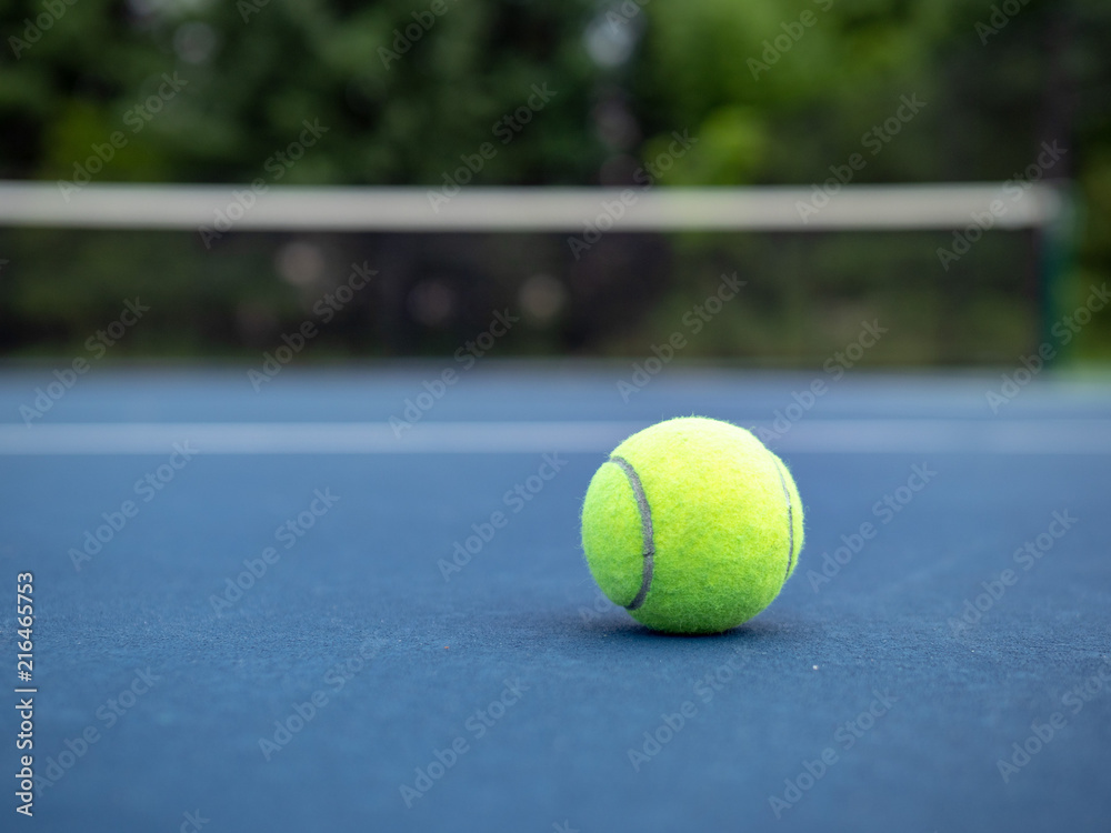 Grounded tennis ball sitting on ground with court net in background 