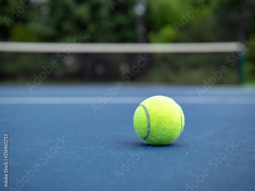 Grounded tennis ball sitting on ground with court net in background  © David Tran