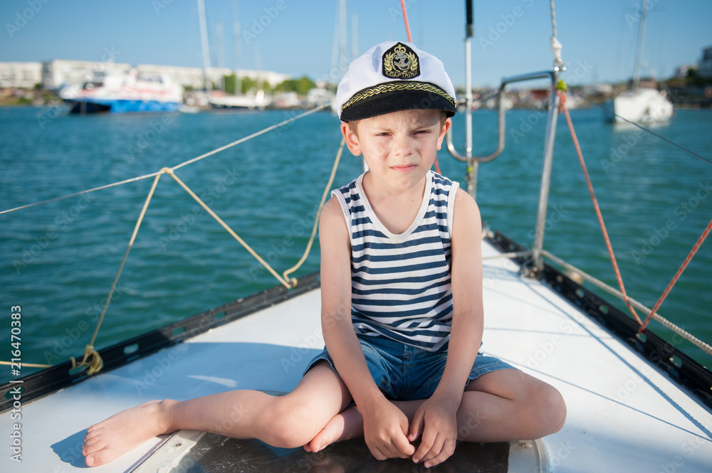 serious kid in captain hat sitting on luxury yacht board in sea port in summer