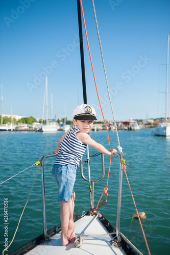 cute kid in captain hat standing on white yacht board in sea bay in summer