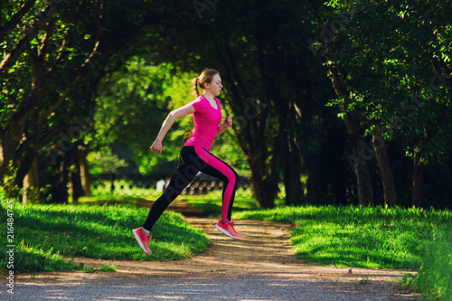 Girl in pink sportswear doing racing jump at green park in summer morning. Healthy lifestyle sport concept.