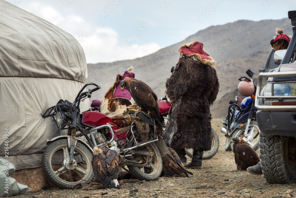 Mongolia,Traditional Golden Eagle Festival, Behind The Scenes Of The Event: Group Of Nomadic Hunters Watching The Competition, Standing Next To Their Vintage Motorcycles And Golden Eagles.