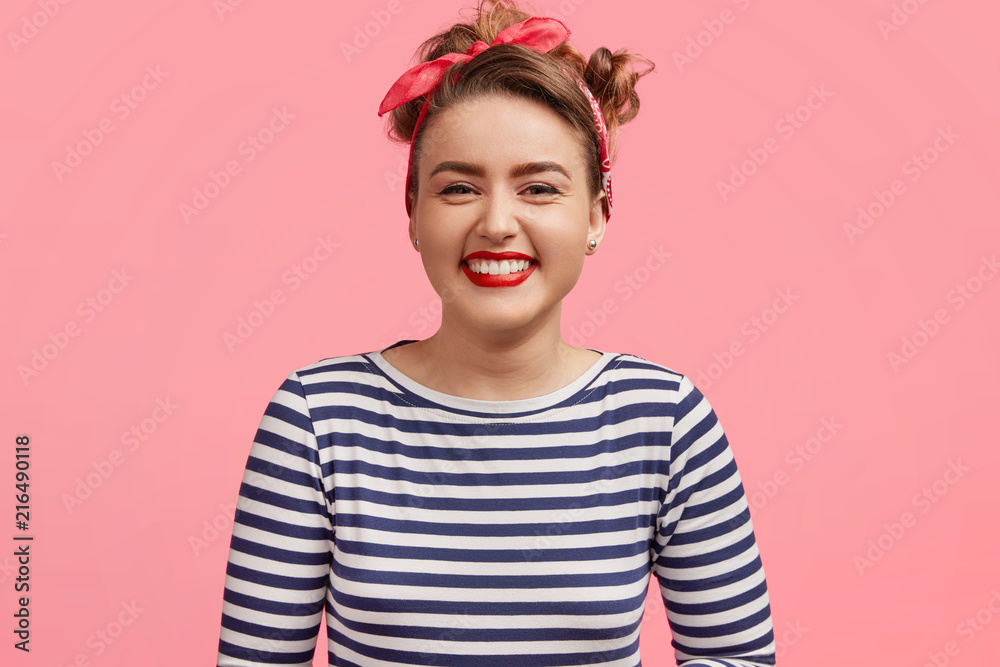 People and happiness concept. Adorable young smiling female dressed in sailor sweater, being pleased by pleasant story, stands against pink background. Joyful pinup girl expresses positiveness People and happiness concept. Adorable young smiling female dressed in sailor sweater, being pleased by pleasant story, stands against pink background. Joyful pinup girl expresses positiveness