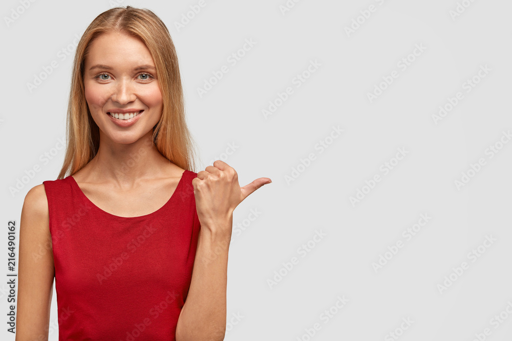 Horizontal shot of pretty happy slim woman has gentle smile, appealing look, points with thumb aside, dressed in red t shirt, happy to meet with talented musician at party. Advertisement concept Horizontal shot of pretty happy slim woman has gentle smile, appealing look, points with thumb aside, dressed in red t shirt, happy to meet with talented musician at party. Advertisement concept