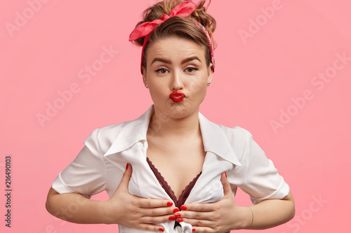 Fashion, beauty concept. Attractive young European female feels proud of her breast, keeps red lips rounded, has nice make up, healthy skin, wears stylish clothes, isolated over pink background. photo