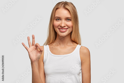 Attractive Caucasian female with long hair, satisfied expression, shows okay sign, feels glad after meeting with handsome guy, isolated over white background. Human facial expressions, body language