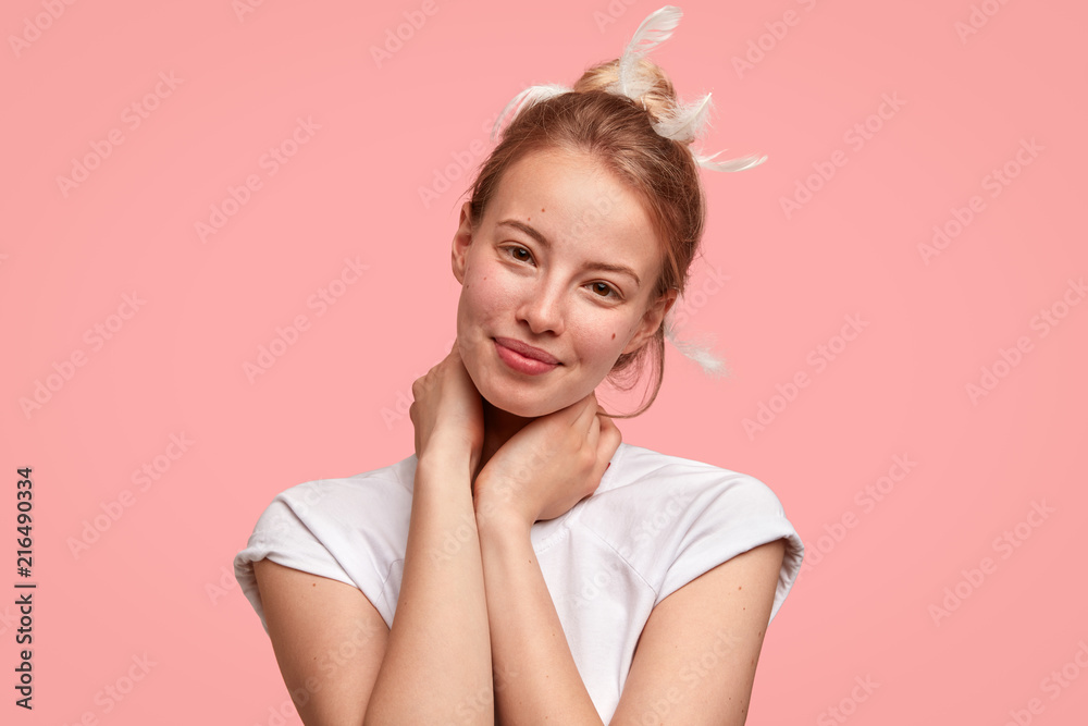 Isolated shot of beautiful female with healthy skin, keeps hands on neck, smiles positively, has appealing look, feathers in hair, wears casual white t shirt, hears something good. Sleeping concept Isolated shot of beautiful female with healthy skin, keeps hands on neck, smiles positively, has appealing look, feathers in hair, wears casual white t shirt, hears something good. Sleeping concept