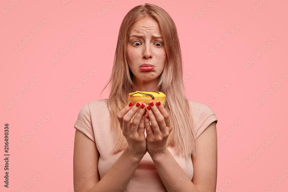 Discontent blonde female holds sweet doughnut, looks with temptation, wants to eat, but keeps to diet, dressed in casual t shirt, isolated over pink background. People and junk food concept. Discontent blonde female holds sweet doughnut, looks with temptation, wants to eat, but keeps to diet, dressed in casual t shirt, isolated over pink background. People and junk food concept.
