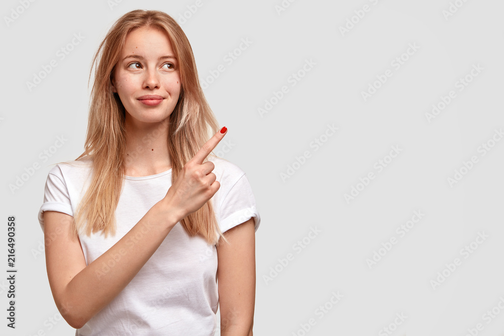 Beautiful pleasant looking blonde female points upwards aside with fore finger, has pleased facial expression, wears casual white t shirt, indicates at blank free space for your advertising content Beautiful pleasant looking blonde female points upwards aside with fore finger, has pleased facial expression, wears casual white t shirt, indicates at blank free space for your advertising content
