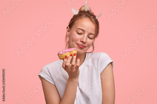 People and temptation concept. Attractive young European female looks at sweet doughnut  going to eat for breakfast  fonds of junk food  dressed casually  isolated over pink studio background