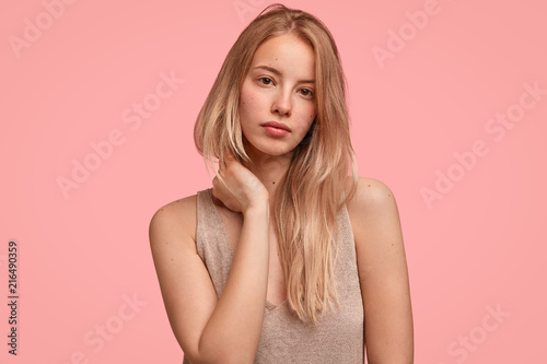 Portrait of good looking blonde female touches neck with hands, looks mysteriously and seriously at camera, has pure skin, going to have outdoor walk, models in studio against pink background