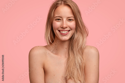 Indoor shot of cheerful Caucasian female with friendly expression and charming smile, stands half nude against pink background, has white even teeth and clean soft skin. Positiveness concept