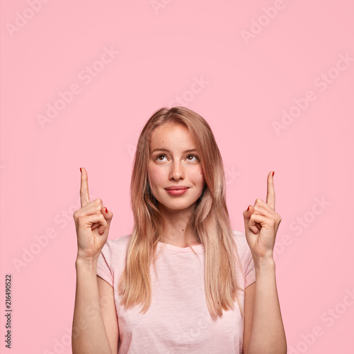 Cute blonde female with long hair, indicates with two fore fingers upwards, s...
