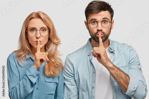 Portrait of mute pretty young female and male makes hush sign, ask to be quiet, keep fore fingers on lips, stand closely, isolated over white background. People, conspiracy and secrecy concept