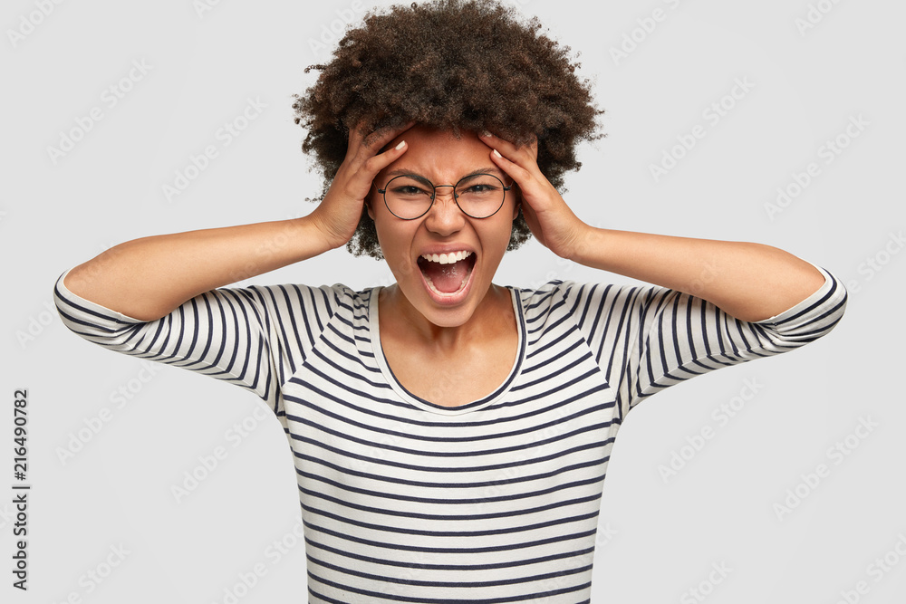 Portrait of depressed displeased dark skinned female with Afro haircut, exclaims with negative feelings, keeps hands on temples, feels desperate, dressed in striped jacket, isolated over white wall Portrait of depressed displeased dark skinned female with Afro haircut, exclaims with negative feelings, keeps hands on temples, feels desperate, dressed in striped jacket, isolated over white wall