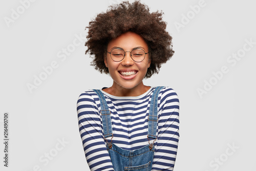 Joyful lovely dark skinned female with pleased expression, has broad smile, closes eyes in happiness, wears fashionable overalls, expresses positive emotions, isolated over white background. © wayhome.studio 